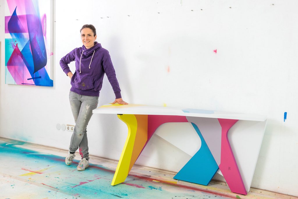 Artist MadC standing with her designed folding table titled Never Stop Moving, against a white wall and a floor covered in pastel paint.