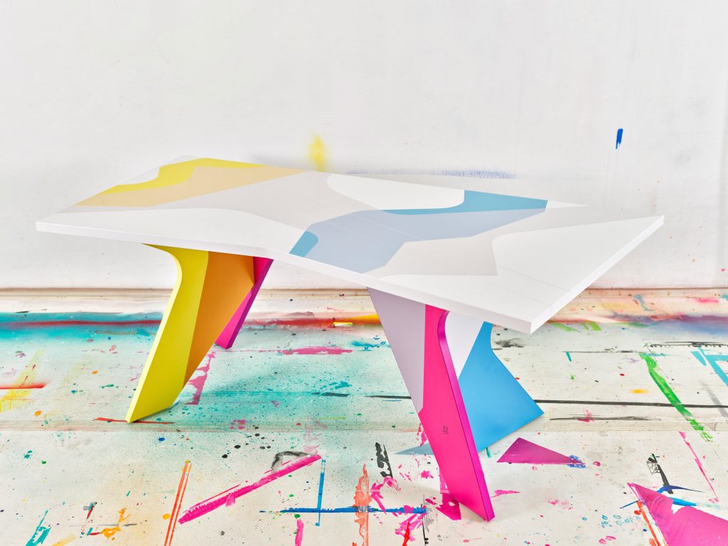 The folding table with brightly colored irregularly shaped panel legs and swathes of transparent color across the top, designed by MadC. The floor has spatters of brightly colored paint.