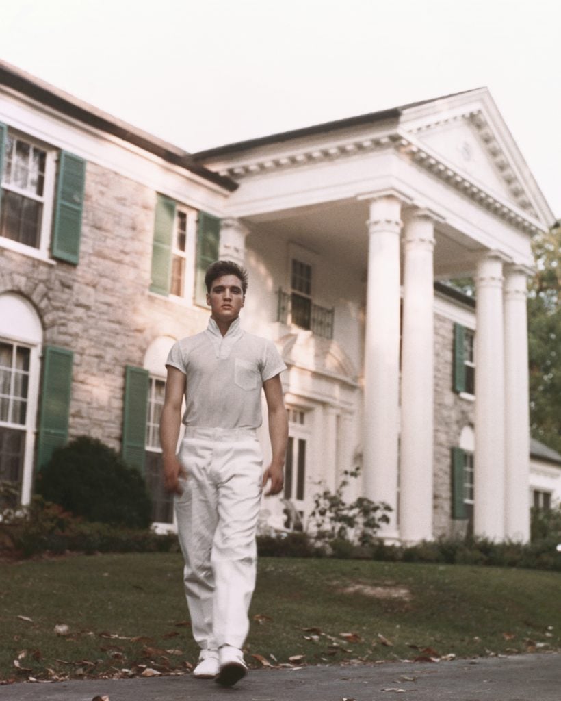 A man, singer Elvis Presley, standing outside a grand mansion