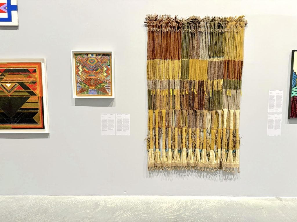 A small abstract painting next to a hanging abstract weaving