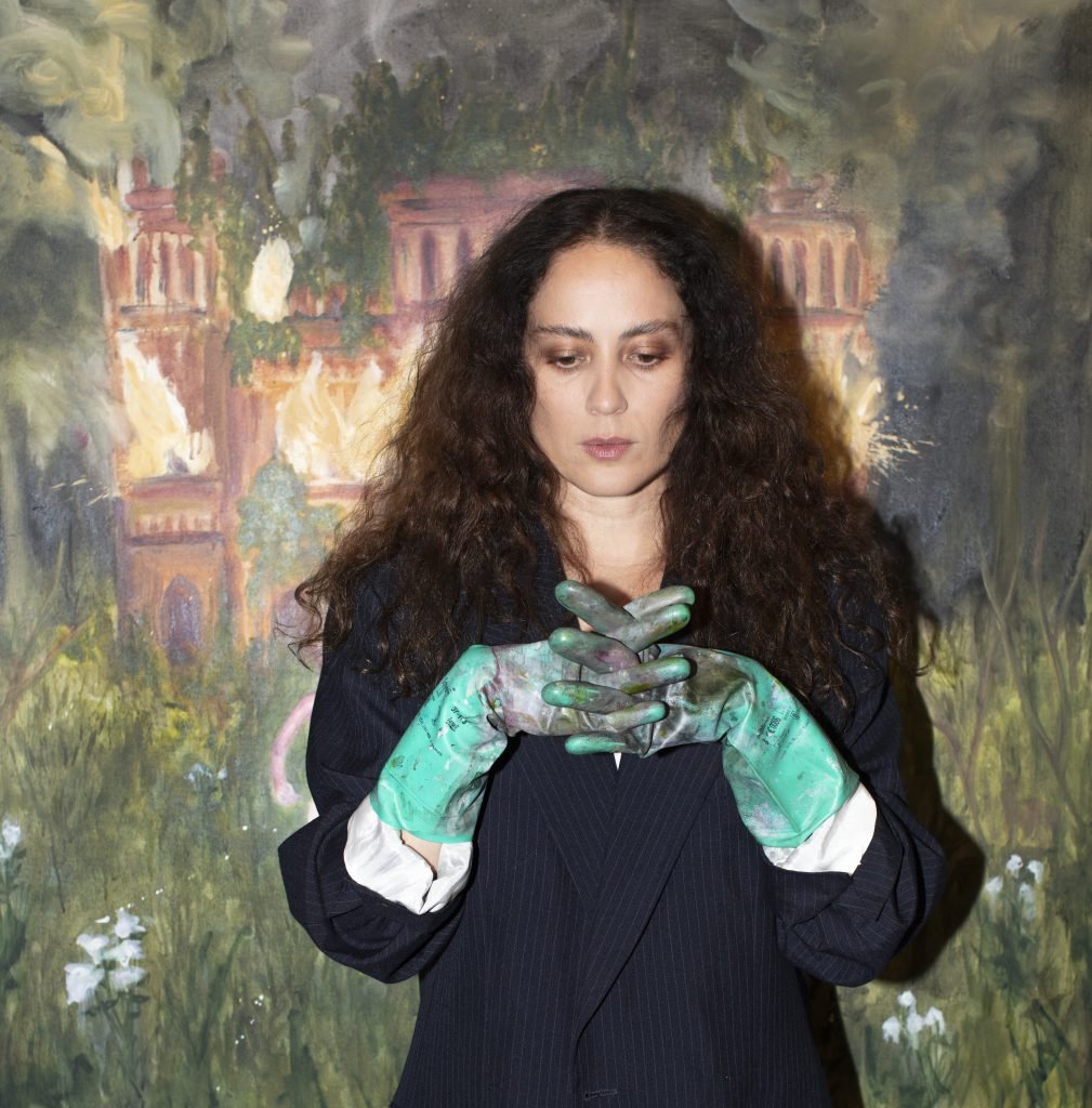 a woman with long curly brown hair stands in front of a painting. she is wearing green latex gloves covered in paint.