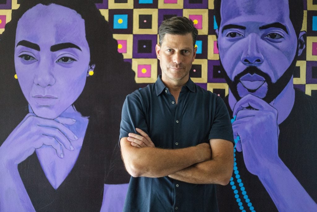 Mestre Projects Founder and CEO Jose Mestre wearing a navy blue button down tee shirt in front of a painting of two portraits done in shades of purple.