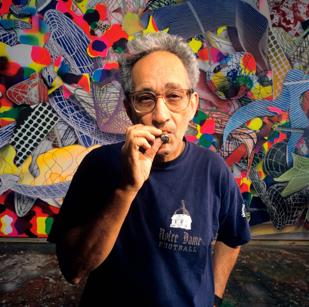 Frank Stella chomping a cuban cigar in front of a colorful painting