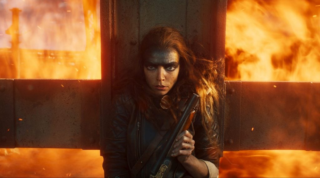 A woman holding a gun standing against a metal pole while a fire rages behind her
