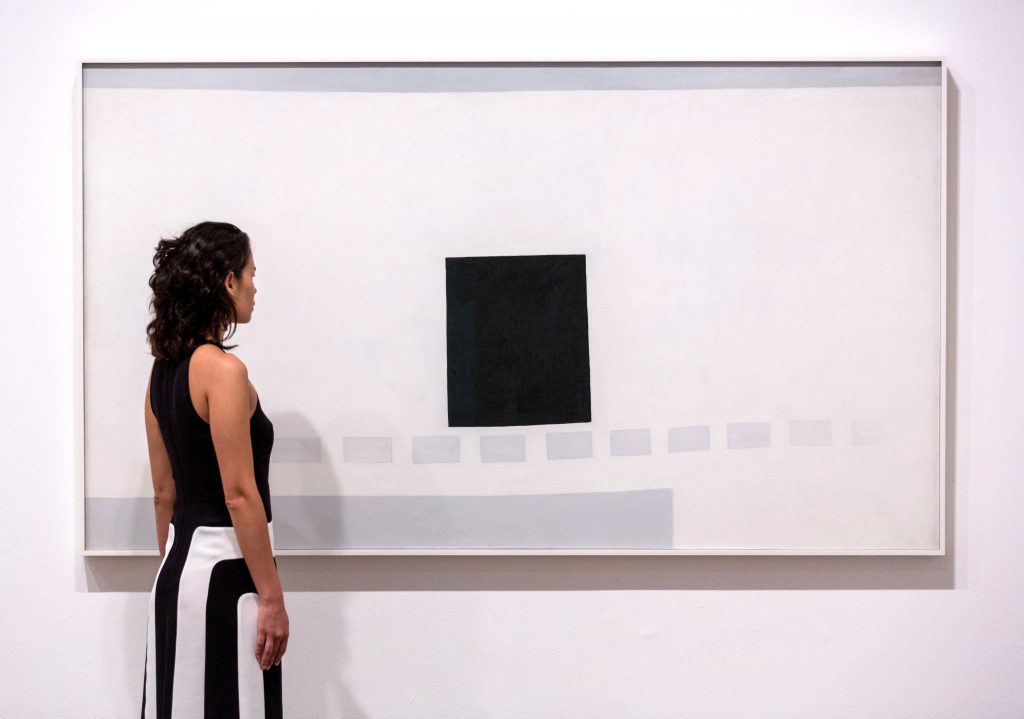 A woman looking at a painting of a black square on a white background, with several smaller grey squares beneath it.
