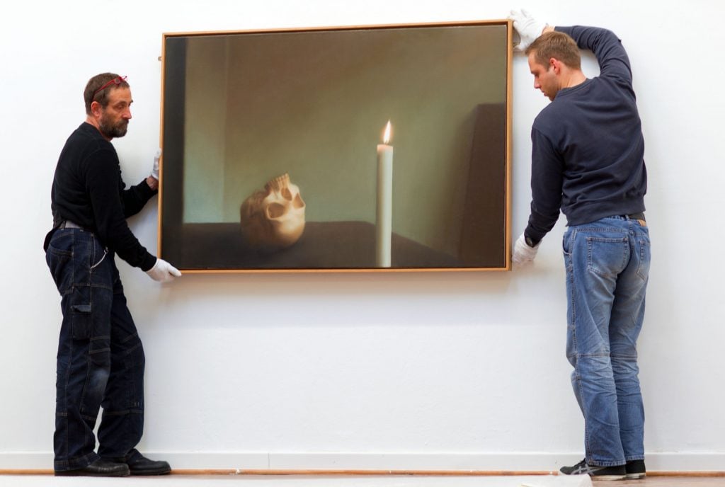 two men hanging a painting by gerhard richter of a candle and a skull