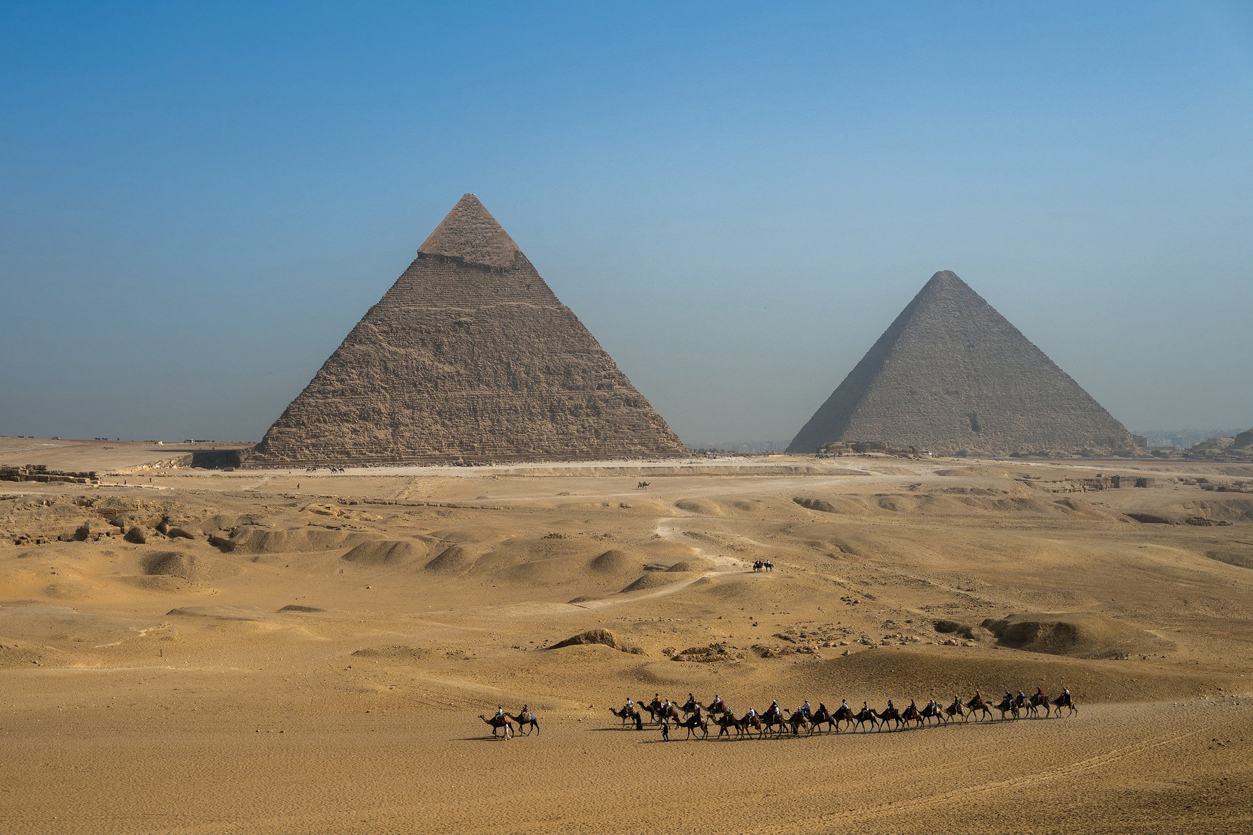 two ancient Egyptian pyramids stand against a blue sky. Tourists on camels walk across the desert in the foreground.