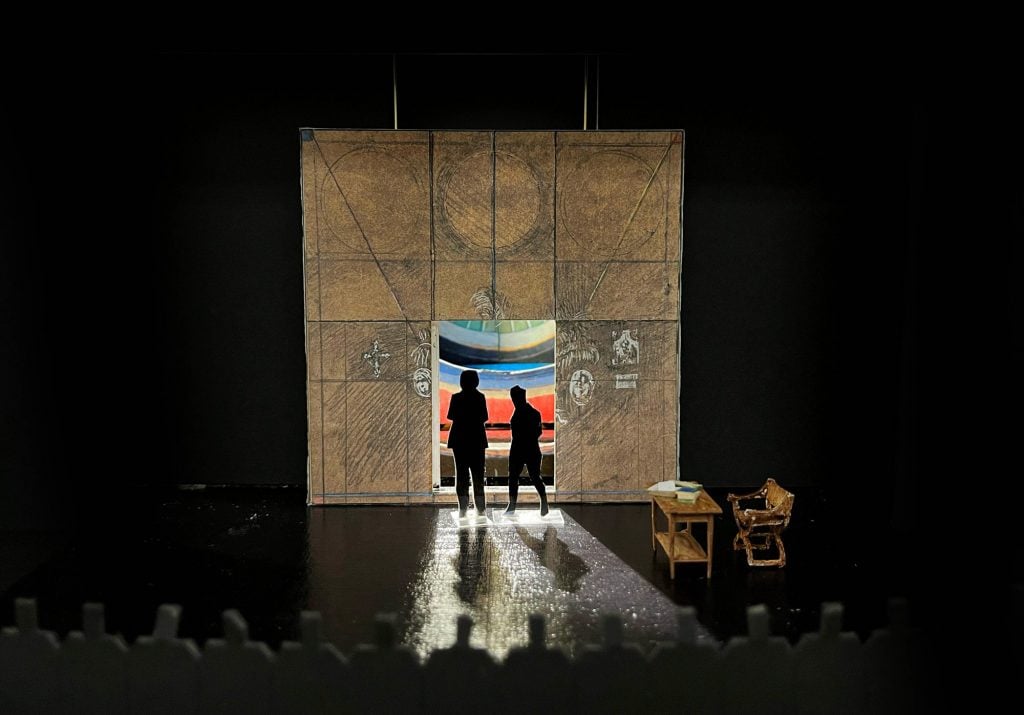 A mock-up of a theater set showing a square backdrop with a doorway cut into in, with two cut-out figures standing in its light