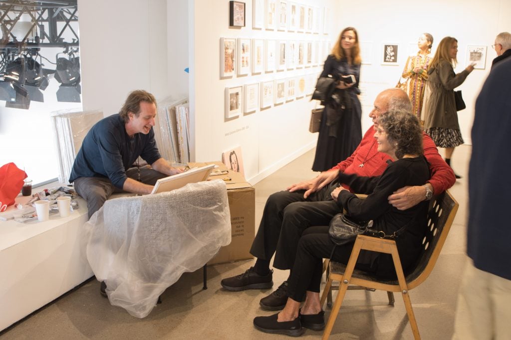 An elderly couple sitting in a bench across from an artist, Joel Mesler, who is working at a canvas.