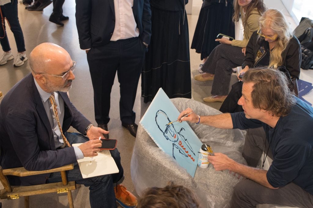 A man sitting across from another man, artist Joel Mesler, who is painting his portrait on a pale blue canvas