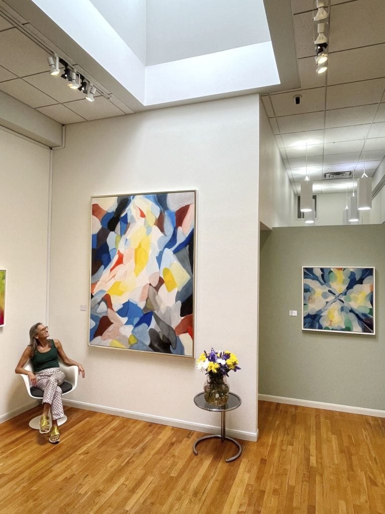 Gallerist Jody Klotz sitting next to a colorful abstract painting inside the exhibition of works by Alice Baber.