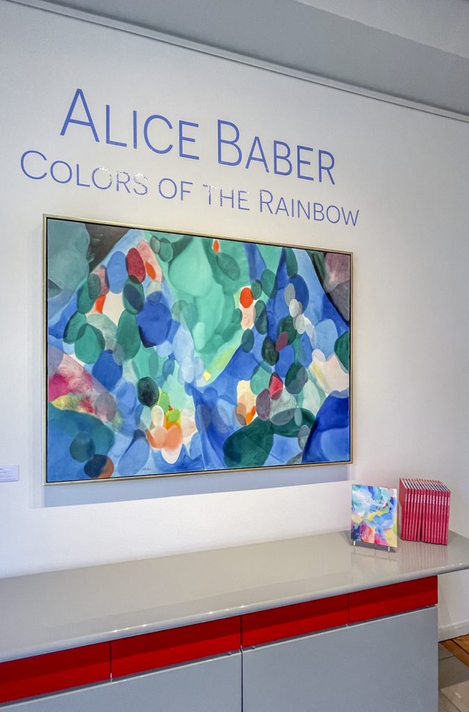 Installation view of an abstract painting with the exhibition title "Alice Baber: Colors of the Rainbow" in vinyl on the wall above, and a row of exhibition catalogues off to the side.