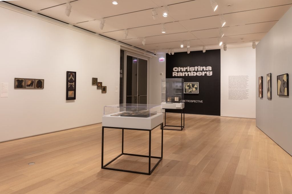 View inside the first gallery space of the Christina Ramberg retrospective at the Art Institute of Chicago, with paintings installed on the wall, a black feature wall with white vinyl text with the artists name, and two standalone vitrines with flat works.