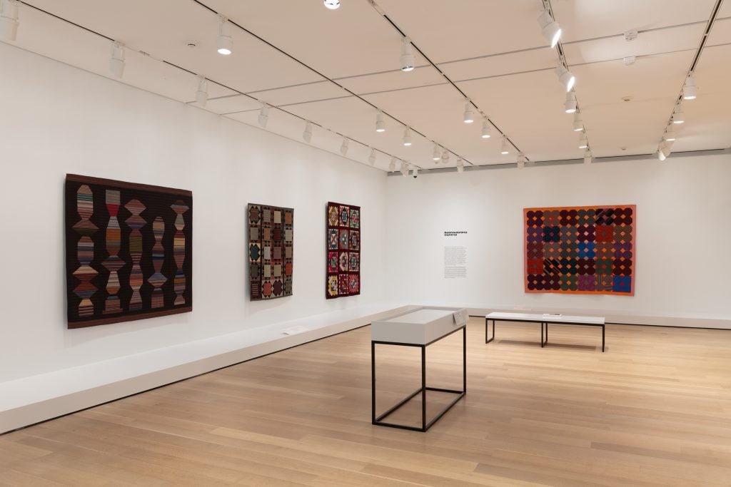 Installation view of "Christina Ramberg: A Retrospective" (2024), featuring four of her quilts installed along the white gallery wall with two vitrines (contents not viewable) in the center of the room.