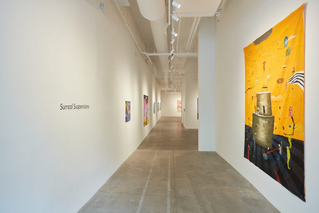 Looking down the entrance hall of the solo show of works by Johan Galue at Unix Gallery, with the name of the show, Surreal Suspensions, on the left wall and a bright, tapestry like painting on the right wall.