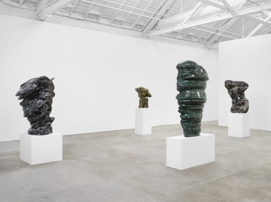 Installation view of four abstract bronze sculptures on pedestals by Tony Cragg inside a white gallery space.