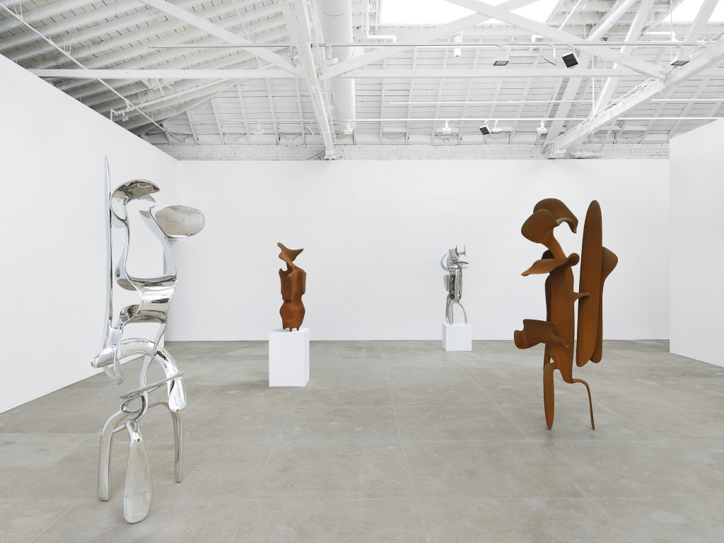 A series of stainless steel and corten steel abstract sculptures by artist Tony Cragg in a white gallery room space.