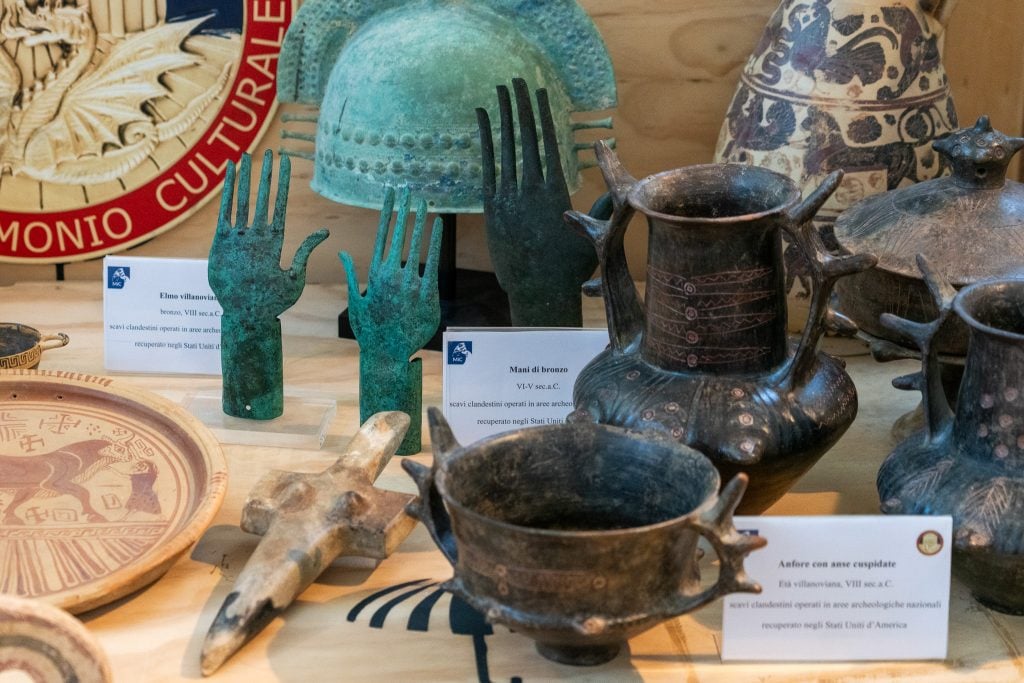 A table filled with ancient artifacts including vessels, sculptures, and a helmet