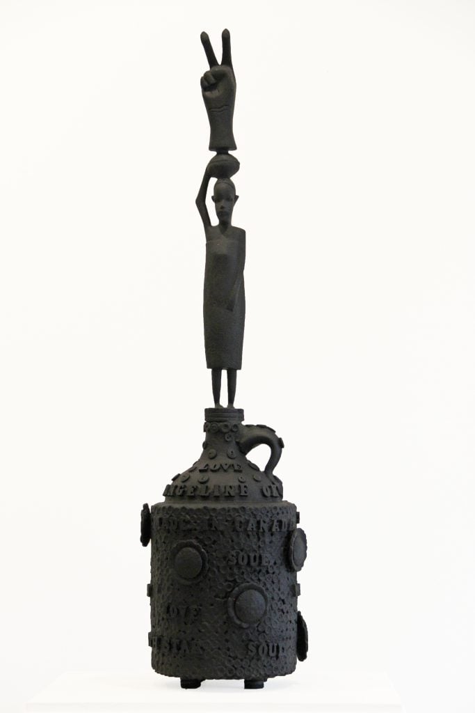 A dark colored assemblage sculpture of a jug with a figure on top carrying a sculpture of a hand in a peace sign on its head,, by Jan Wade.