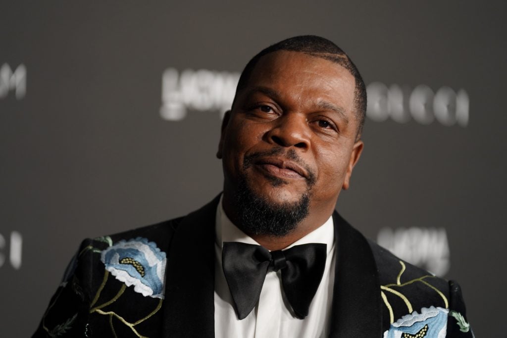 A man, artist Kehinde Wiley, in a tuxedo against a black graphic background