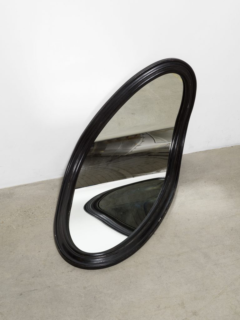 photograph of an oval warped black frame mirror laying against the florr and wall