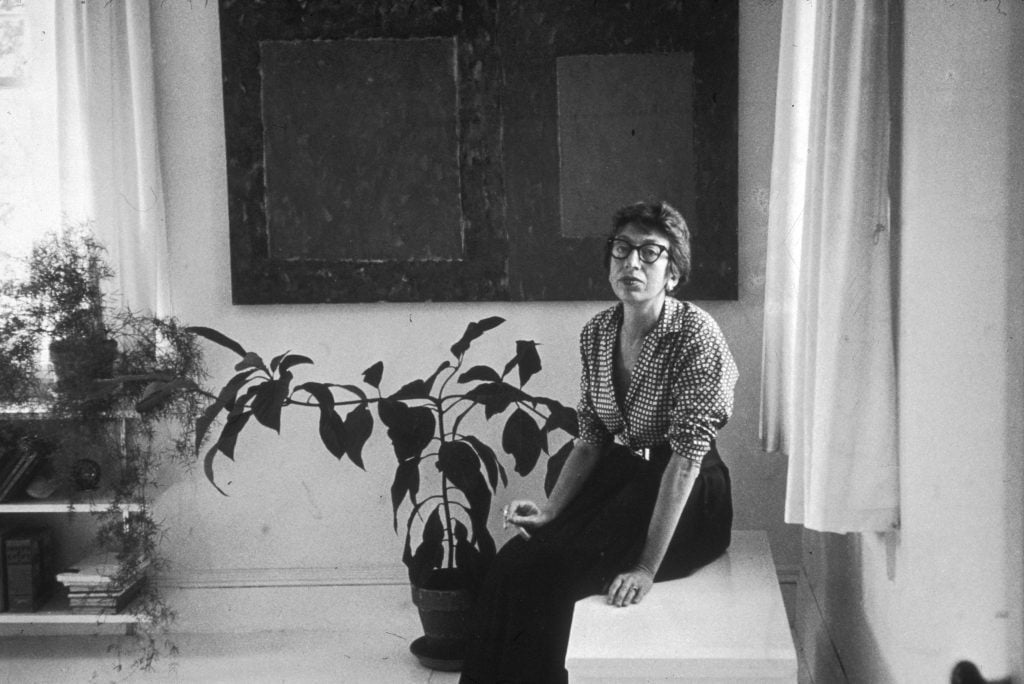 A woman sitting on a window ledge in front of a plant and a painting hanging on a wall.