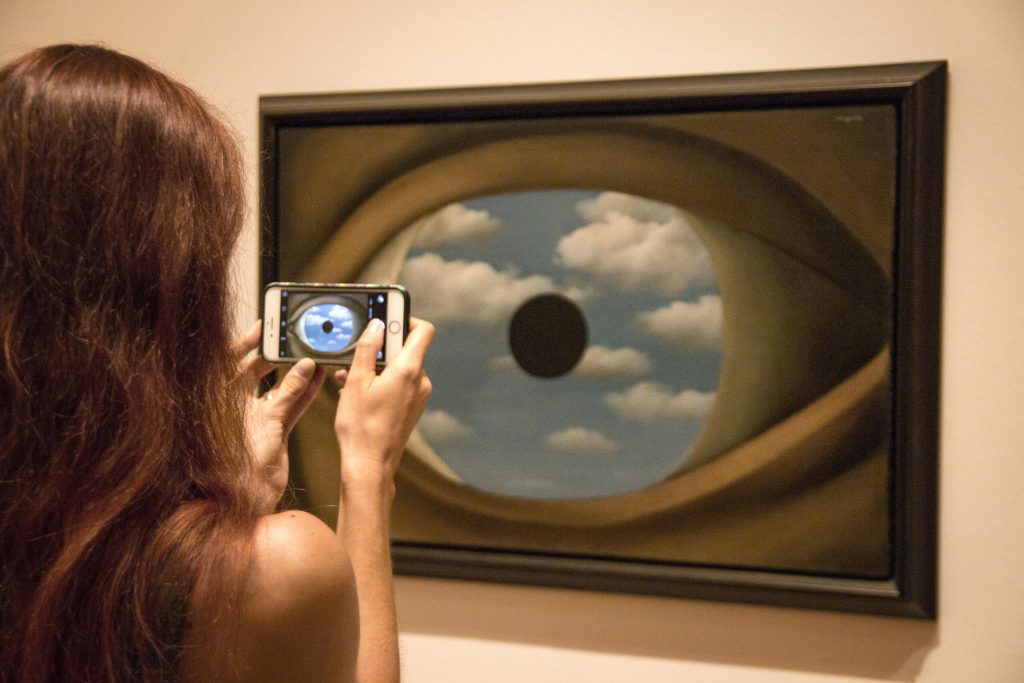 A woman with the smartphone is taking a picture of a painting of huge human eye, with its iris replaced by a blue sky