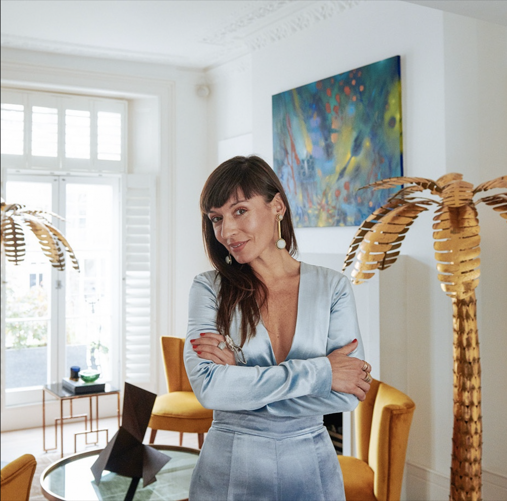 Portrait photograph of Maria Vega, founder of Ministry of Nomads, wearing silver blue shirt and pants in a dining room setting with gold palm tree sculptures.