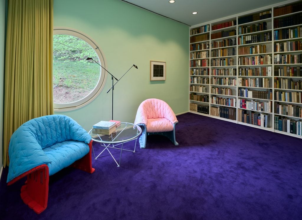 A reading room with mint green walls and purple carpeting. A bookshelf is built into one wall, alongside a blue and pink chair, and a coffee table