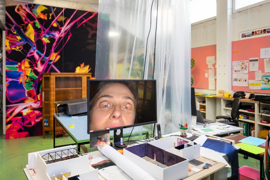 photograph of a miniature model of a gallery on a desk with a computer screen showing a woman with eyelids pulled up