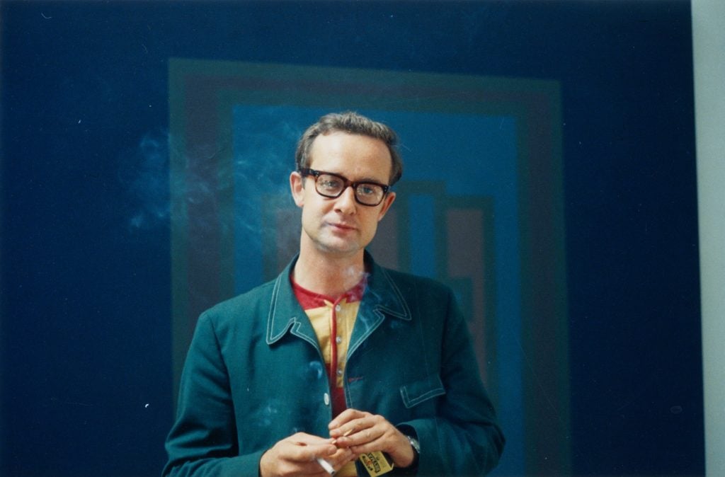 A bespectacled man smoking in front of a blue painting