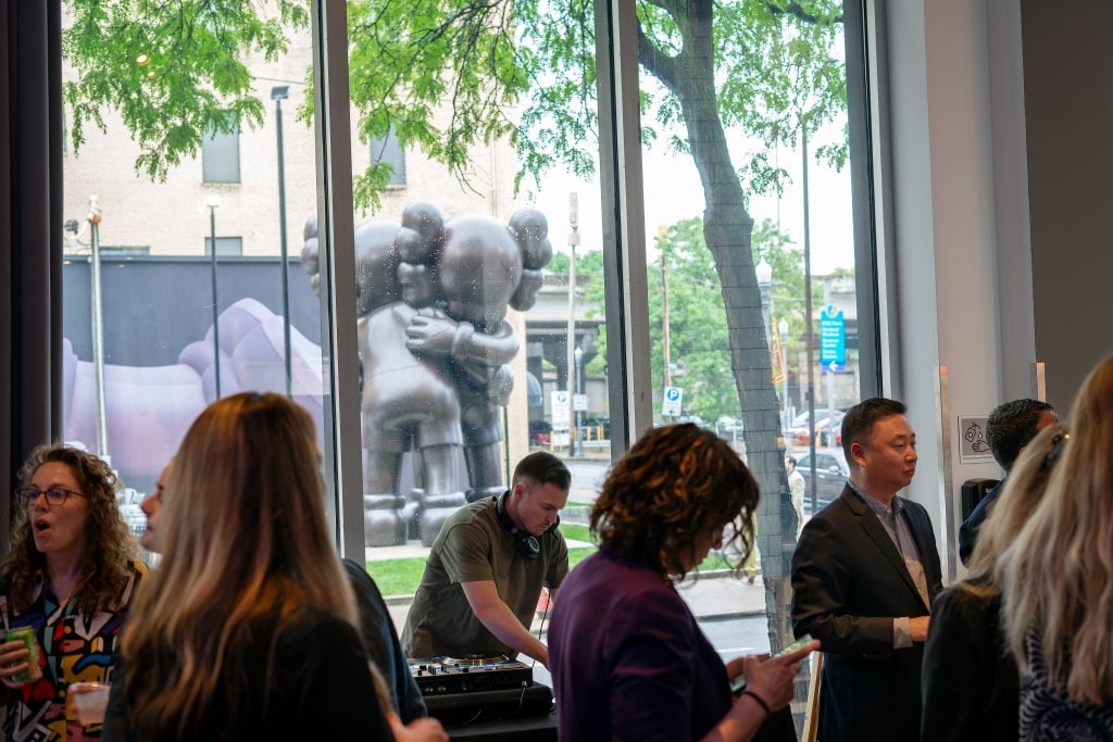 A group of people is gathered inside a gallery, some engaged in conversation while others use their phones. A DJ is seen mixing music on a console. Through the large windows, a large outdoor sculpture of two figures embracing is visible. The setting is lively, with natural light coming in through the windows, showcasing a tree and buildings outside.