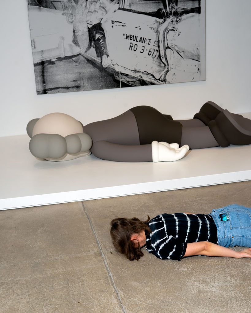 A person lies face down on the floor in front of a large, reclining sculpture of a cartoon-like character. The sculpture, which features rounded shapes and muted colors, is displayed on a white platform. Behind the sculpture is a black-and-white artwork depicting a car crash scene with an ambulance. The person on the floor is wearing a black and white tie-dye shirt and blue jeans. The scene creates a surreal and contemplative atmosphere in the gallery space.