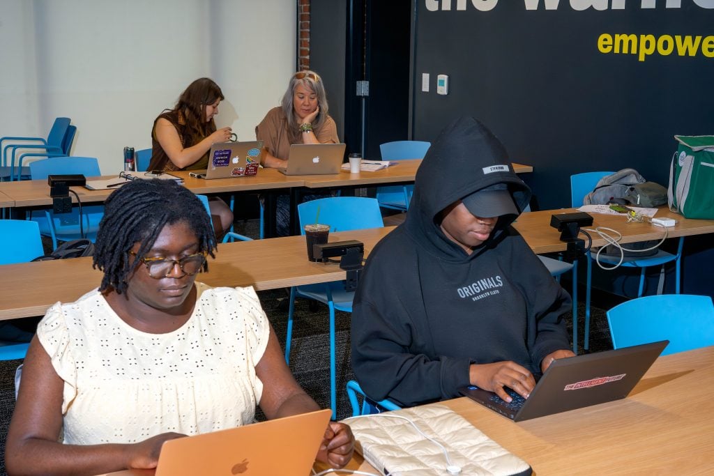 A group of four people are working on laptops in a classroom setting. In the foreground, two individuals are focused on their work; one is wearing glasses and a light-colored top, while the other is wearing a black hoodie and cap. In the background, two other people are also using laptops, with one person wearing sunglasses on their head. The room has blue chairs and wooden tables, with a wall that has the words "the warhol" and "empower" in bold yellow and white letters.
