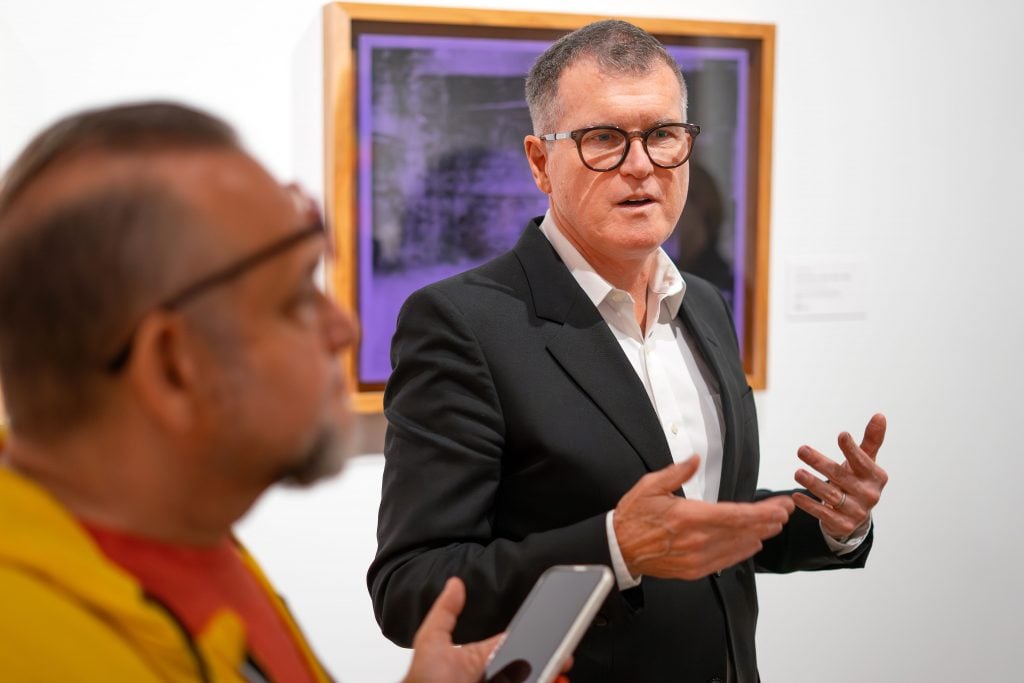 A person wearing glasses and a black suit is speaking, gesturing with their hands. Another individual, partially visible in the foreground, is holding a smartphone. The scene is set in an art gallery, with a purple artwork in a wooden frame on the white wall behind them. The person in the suit appears engaged in conversation, while the background features a blurred figure observing the interaction.