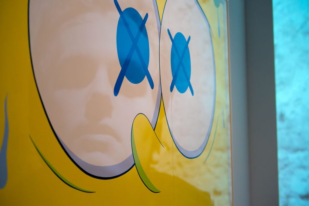 A close-up shot of a colorful artwork featuring a yellow face with large blue eyes, each with an "X" through them. The reflection of a person's face is faintly visible on the surface of the artwork, blending with the illustrated face. The background includes hints of blue and white colors, suggesting a modern, vibrant setting.