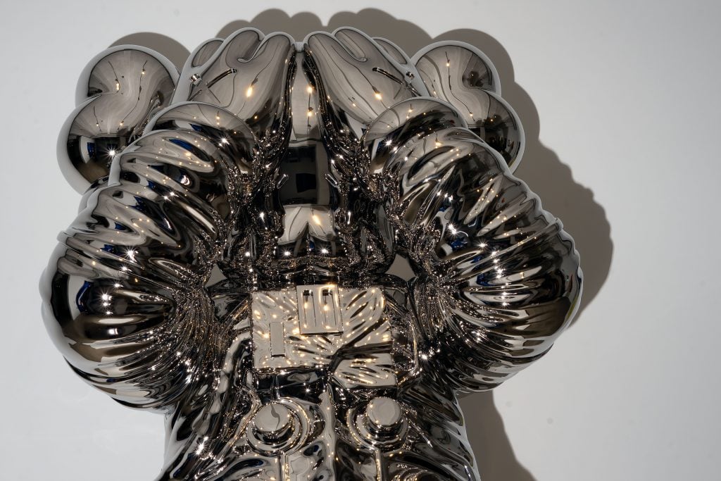 A close-up view of a highly reflective, chrome-colored sculpture depicting a character covering its face with its hands while wearing a space suit. The surface of the sculpture is shiny and polished, reflecting light and surrounding objects. The character has exaggerated, rounded features and intricate detailing, creating a striking visual effect against the plain white background.