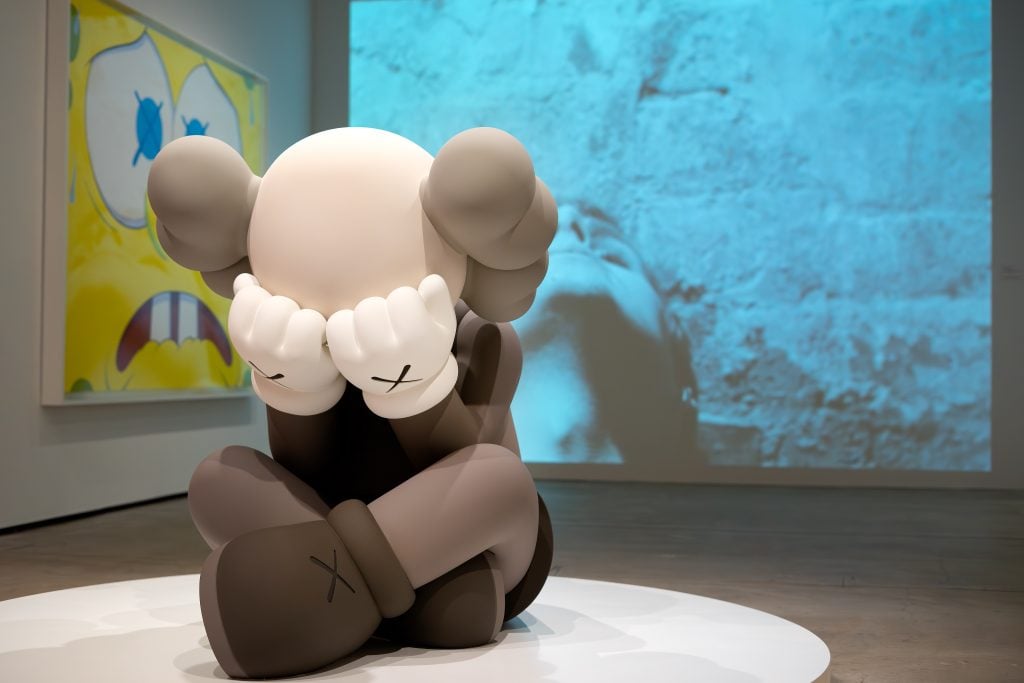 A large, cartoon-like sculpture of a character with a round head and "X" marks on its hands and feet is sitting on a white platform, covering its face with its hands. In the background, a yellow artwork with blue "X" eyes and a large projection of a person’s face against a blue wall are visible. The scene is set in a modern gallery space, highlighting the playful yet melancholic expression of the sculpture.