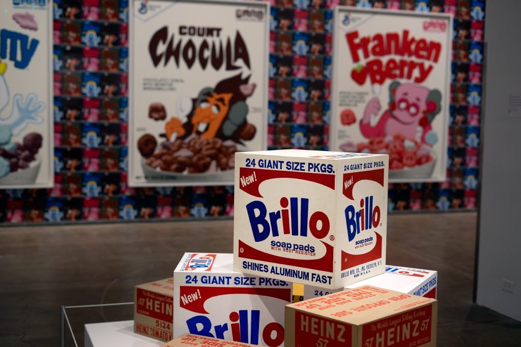 An art installation featuring large boxes resembling Brillo soap pad packaging stacked on a white platform. In the background, there are oversized cereal box artworks for Count Chocula and Franken Berry, with a colorful, patterned wall featuring cartoon characters. The scene captures a playful and nostalgic blend of pop art elements in a gallery setting.