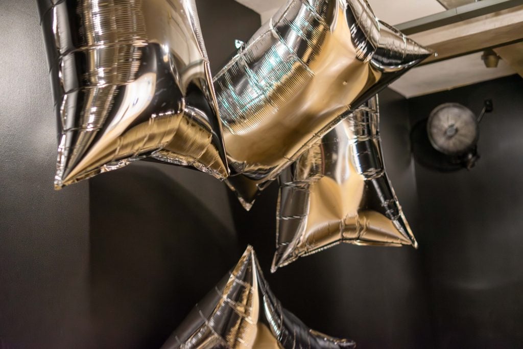 Several silver, reflective balloons shaped like stars are floating in a dark room. The balloons reflect light and their surroundings, creating a shimmering effect. In the background, a ventilation fan is visible on the wall, contributing to the industrial ambiance of the space.