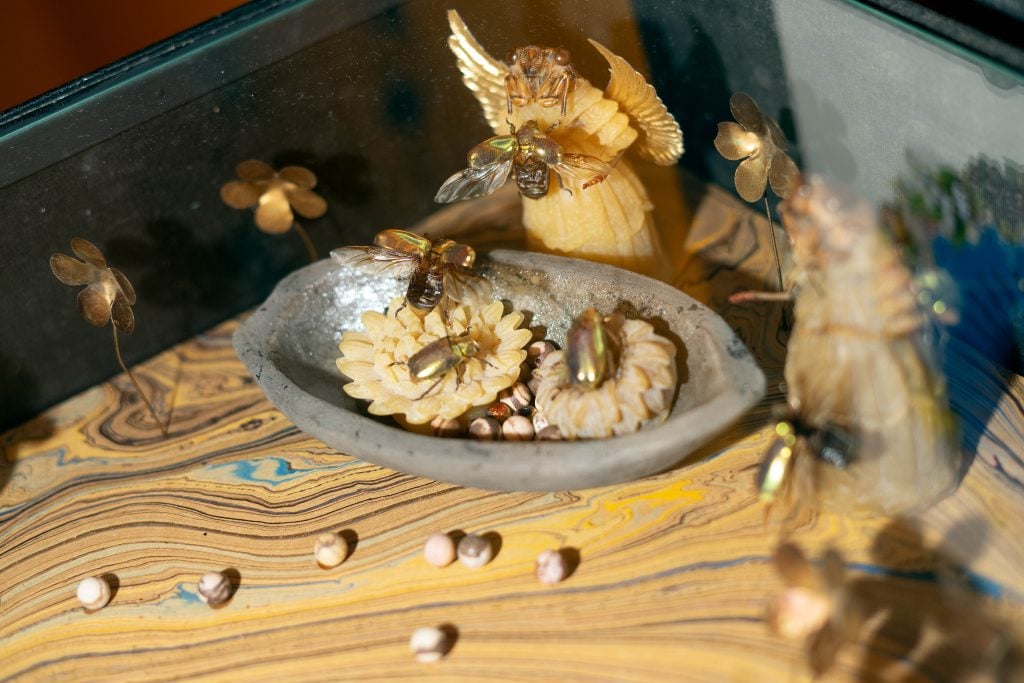 A close-up of a decorative display featuring golden-colored insect specimens arranged on intricately carved flowers within a stone bowl. The background includes small metallic flowers and angel-like figures, all set on a swirling, marbled surface.