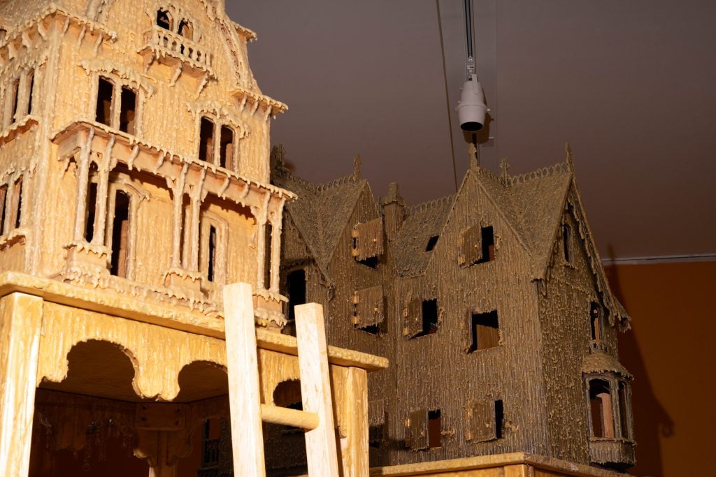 Close-up of a detailed model of a large, gothic-style house made from wax or a similar material. The structure features intricate architectural details, multiple windows, and balconies, with a wooden ladder leaning against it. Another similar house model is visible in the background. The models are displayed indoors.