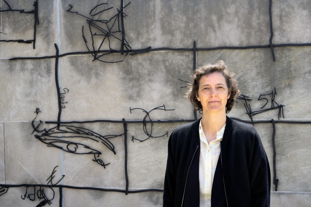 A woman stands in front of an artwork consisting of tangled, black metal lines on a gray stone background. She has a thoughtful expression, short brown hair, and is wearing a dark jacket over a white collared shirt.