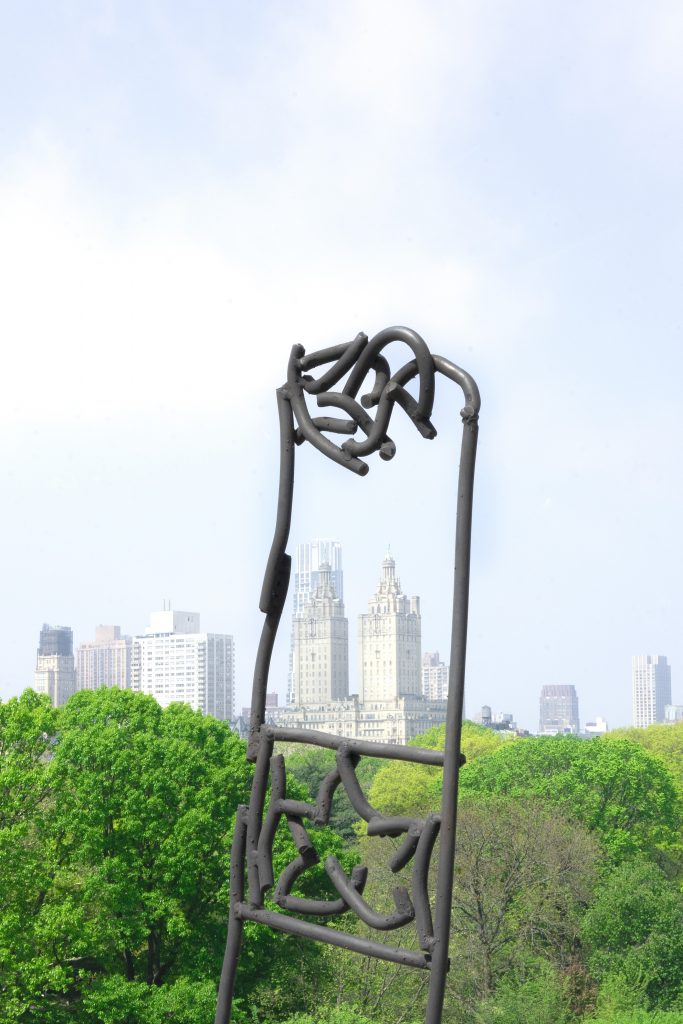 A towering abstract metal sculpture set against a backdrop of Central Park and the iconic New York City skyline. The sculpture features tangled loops and stands prominently over a lush, green landscape.