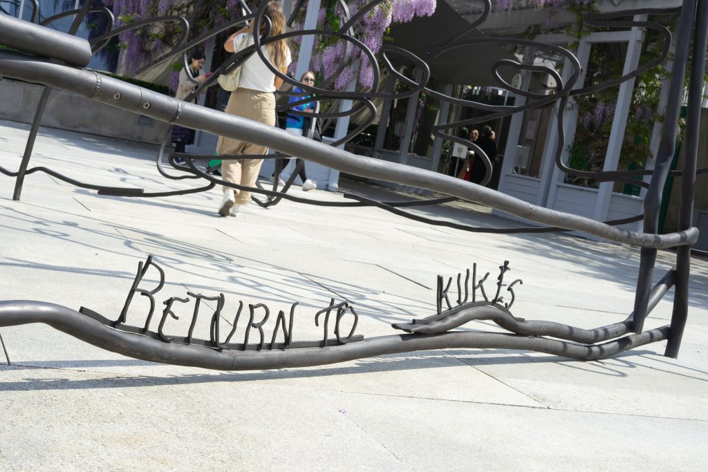 A part of a sculptural installation featuring curving metal rods and the words "Return to Kukës" welded onto a pipe, set on a light concrete ground. In the background, people are walking around the area, which is decorated with purple flowering trees.