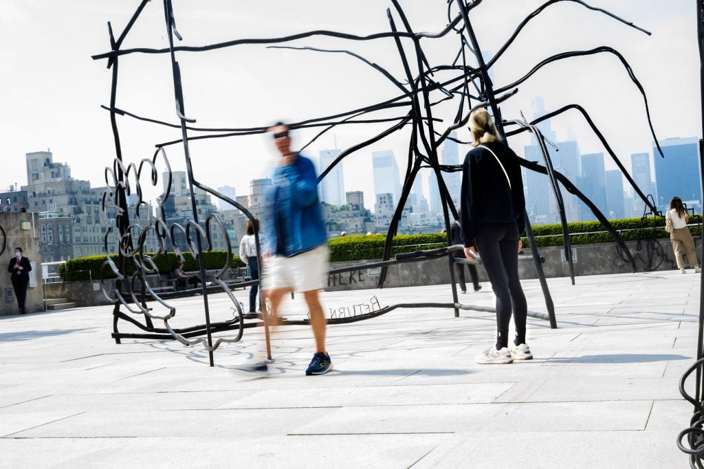 A metal sculpture by artist Petrit Halilaj with complex, intertwined lines stands in an outdoor setting. People are walking and interacting around the sculpture, with blurred motion indicating their movement. The city skyline forms the background, adding an urban context to the scene.