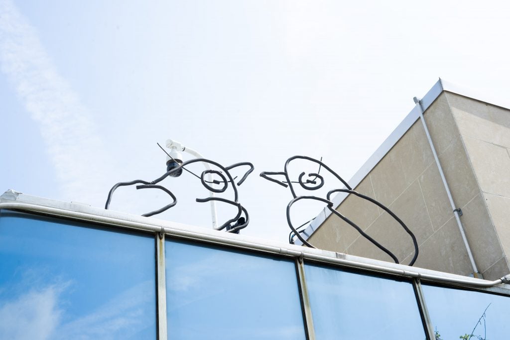 A whimsical metal sculpture depicting two bird figures with circular elements, mounted atop a glass building against a clear blue sky. The sculpture's playful and airy design contrasts with the solid, architectural lines of the building.
