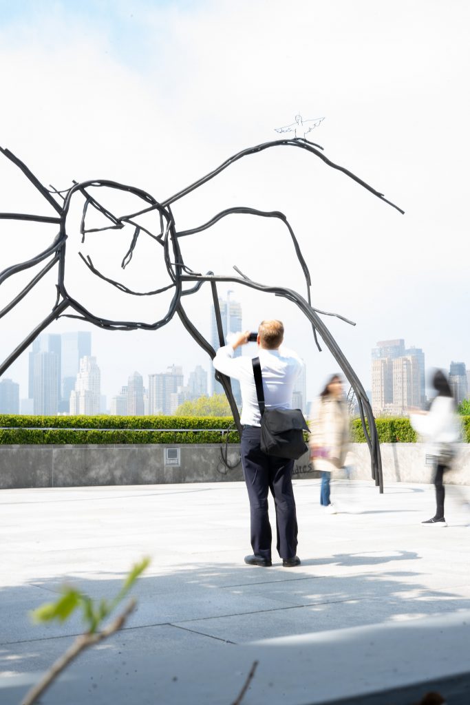 A man in business attire, with a white shirt and suspenders, stands observing an intricate metal sculpture outdoors. The background features blurred figures in motion and a city skyline, creating a dynamic urban scene.