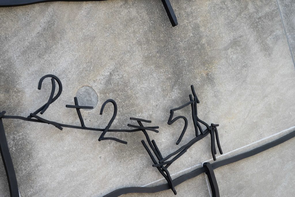Close-up of a section of a metal sculpture displaying abstract numerical and alphabetical shapes in a flowing script on a textured gray concrete background.