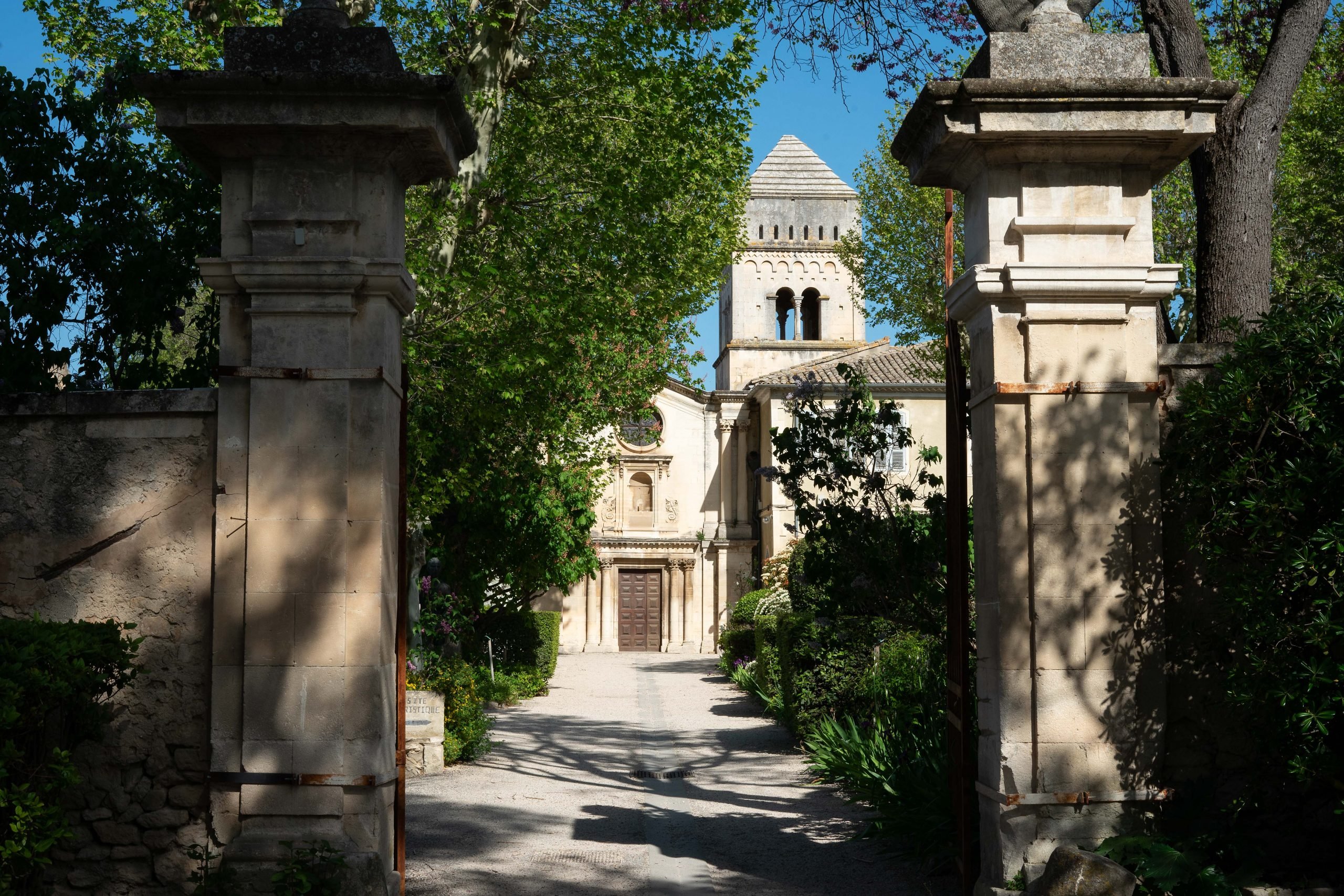 An open gateway leading to a historic building, the Monastery Saint-Paul de Mausole, with a classical facade and a bell tower, surrounded by lush greenery and a gravel path.